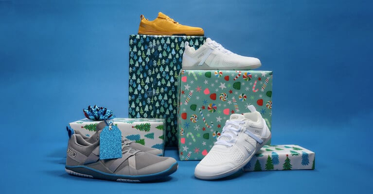 Festive display featuring a Frost Grey Forza Trainer, White Prio, and Gold Nexus Knit on gift boxes with holiday wrapping.