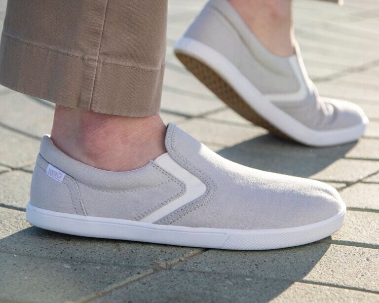 Closeup of men's feet wearing light gray Dillon Canvas Slip-On sneakers while standing on a brick walkway in the sun