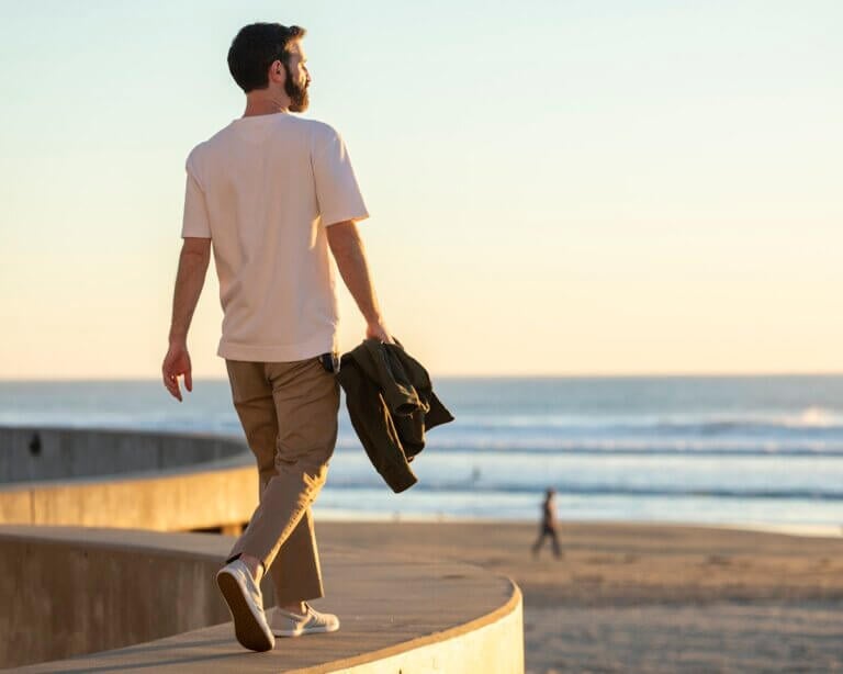 A man wearing light gray Dillon Canvas Slip-On sneakers walking on a concrete wall and looking at the ocean