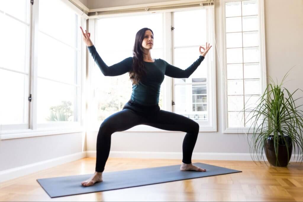 Woman doing goddess pose on a yoga mat at her home in front of well-lit windows