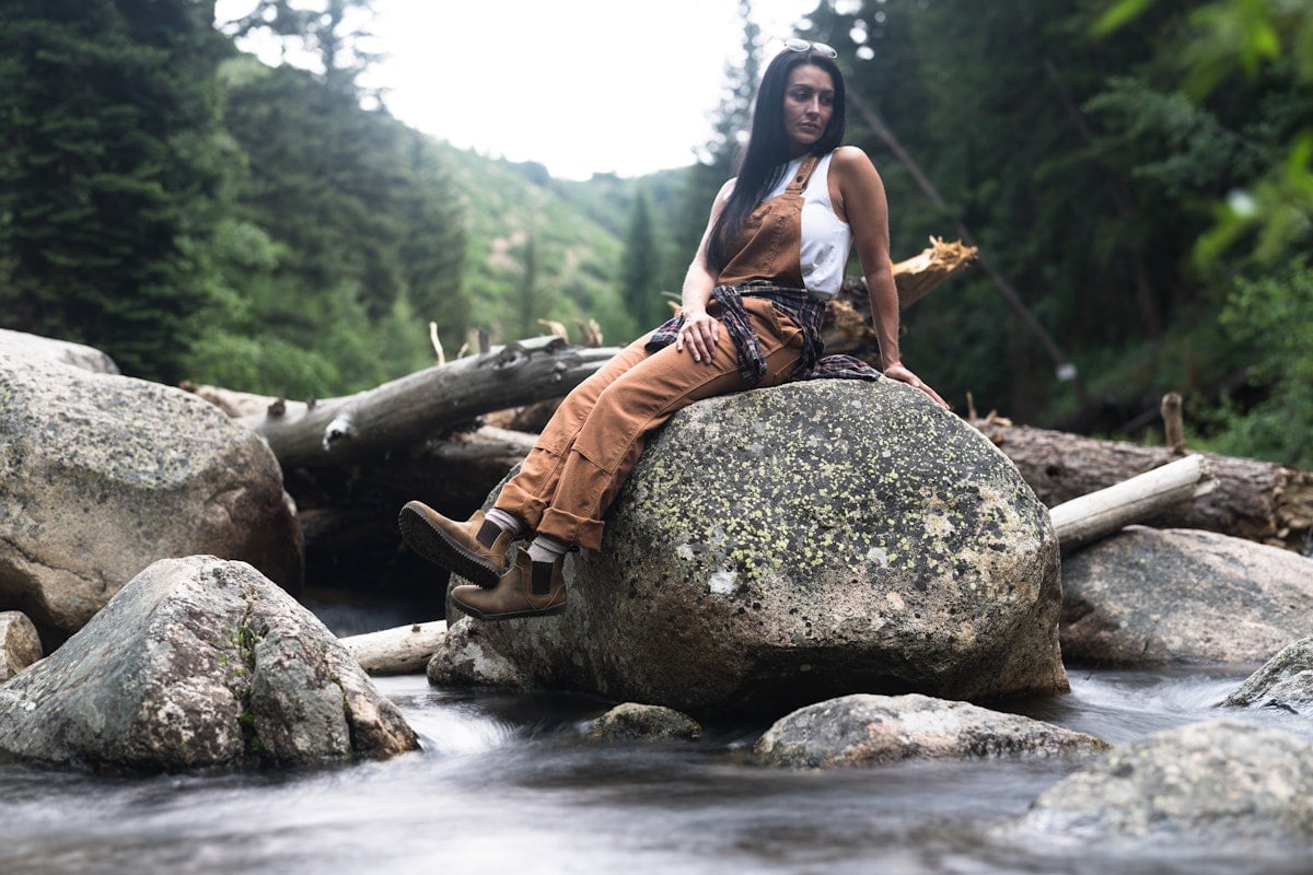 A woman hiker sitting on a large rock by a stream with their Ridgeway Chelsea boots